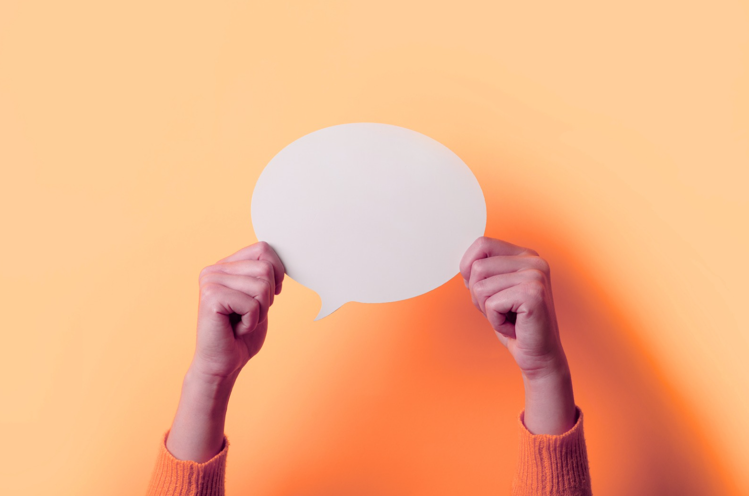 A pair of hands hold up an empty white speech bubble in front of an orange background